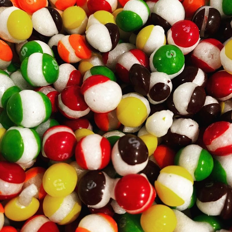 Close-up of colorful freeze-dried Skittles, half-white with red, green, yellow, orange, dark brown, some with 'S' logo.