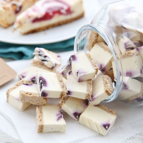 Blueberry swirl cheesecake bites with graham crust spill from a glass jar. Two cheesecake slices on a plate are in the background.