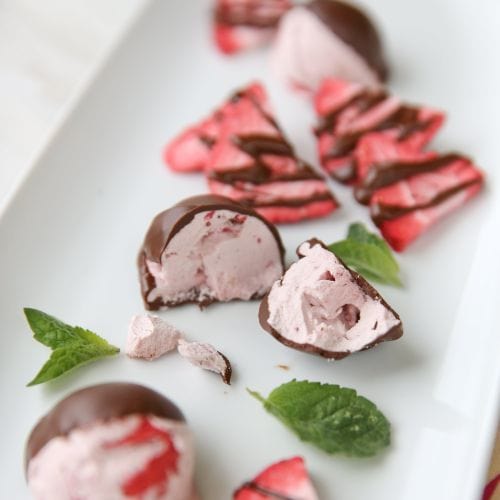 Chocolate-dipped pink freeze-dried fruit bites with fluffy interiors and mint leaves on a white platter.