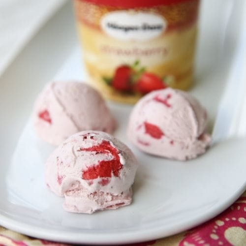 Strawberry ice cream scoops with fruit pieces on a white plate, a blurred Häagen-Dazs strawberry pint behind.