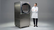 A scientist stands beside a commercial freeze dryer with its door open, revealing trays of food.