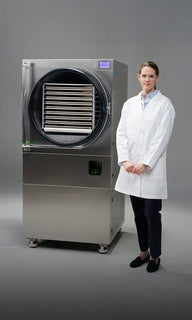 A woman in a white lab coat stands next to a large, stainless-steel freeze dryer.